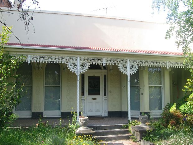 Front exterior of a historic single-storey terrace-style residence with an ornate white cast-iron verandah, decorative lacework frieze, slender columns, tall sash windows, a central entrance door, and a red corrugated metal roof. The pale rendered facade is partly framed by mature garden planting and front steps.