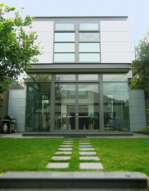 Front exterior view of a contemporary two-storey residential extension or house, with a light grey panelled upper facade, extensive dark-framed glazing and glazed ground-floor doors opening toward a manicured lawn. A stepping-stone path leads to the building, with outdoor equipment visible at the side.