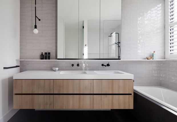 Contemporary bathroom featuring a double wall-mounted timber vanity with integrated white basins, a large mirrored cabinet, pale textured wall tiles, black tapware and fittings, and a partially visible bathtub. Natural light enters through white plantation shutters, creating a clean, refined atmosphere.