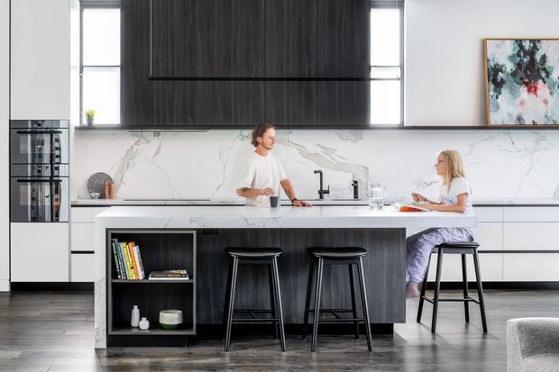 Bright contemporary kitchen with a large dark timber island, integrated open shelving, white marble-look splashback, black appliances and tapware, and seating for four. Two people sit and stand at the island, while tall windows and an abstract artwork add natural light and colour.
