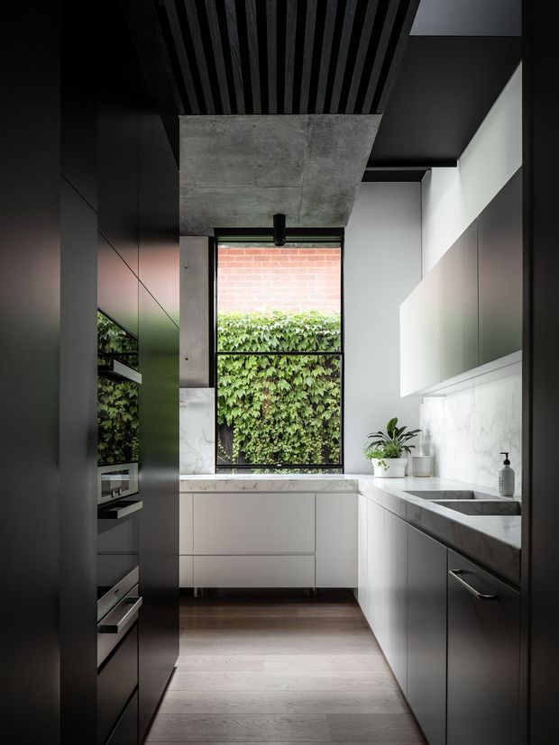 A narrow, sophisticated contemporary kitchen with dark full-height cabinetry, integrated appliances, pale stone benchtops and splashback, and a deep stainless-steel sink. A large black-framed window brings in greenery and daylight, while the ceiling combines exposed concrete, black linear battens, and dark finishes. The restrained palette and clean joinery create a refined, modern atmosphere.