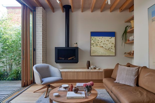 A warm, contemporary living room with exposed timber ceiling beams, a black suspended wood-burning fireplace, and a built-in timber media/storage cabinet. Full-height timber-framed glazing opens to lush planting outside, while a brown leather sofa, upholstered lounge chair, round timber coffee table, and neutral furnishings create a comfortable, refined interior.