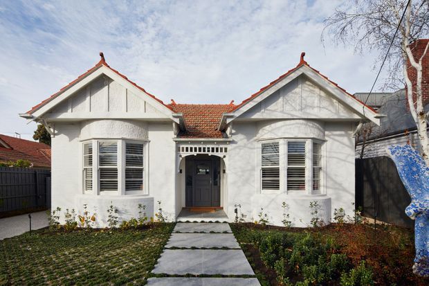 Front exterior view of a symmetrical heritage-style single-storey house with two gabled wings, white rendered walls, decorative gable detailing, red terracotta tiled roofing, timber-framed windows with white shutters, and a central recessed entry. A paved path leads through landscaped front gardens.
