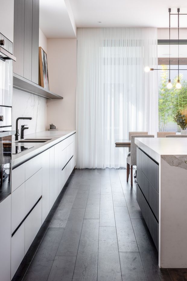 A bright contemporary kitchen viewed along a galley-style layout, with white flat-panel cabinetry, dark integrated appliances and hardware, a pale veined stone backsplash and island benchtop, and dark timber-look flooring. The kitchen opens toward a dining area with upholstered chairs and a large window screened by sheer curtains. Black linear pendant lights hang above the island, while vertical timber-toned cabinetry and open shelving add contrast.