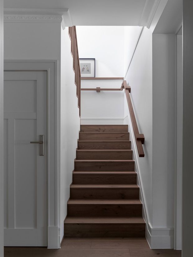 A bright residential stairway viewed from the lower hallway, with dark stained timber stair treads and flooring, white walls, traditional white decorative cornices and door trim, and a simple timber handrail and balustrade. A landing and framed artwork are visible at the top of the stairs.