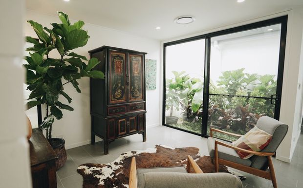 Bright, refined sitting area with a large fiddle-leaf fig, ornate dark timber armoire, mid-century-style upholstered armchair, and cowhide rug. Floor-to-ceiling black-framed glazing opens the room visually to a lush planted courtyard or garden, while recessed lighting and pale surfaces keep the space airy.