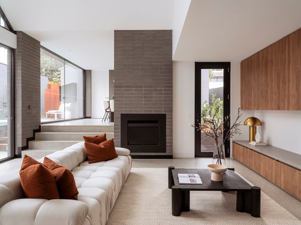A bright contemporary living room with a large white modular sectional sofa, rust-coloured cushions, a dark rectangular coffee table, and a built-in fireplace set into a full-height grey tiled feature wall. Warm timber wall cabinetry lines the right side above a low console, while full-height glazing, stepped flooring, and an adjoining dining area create an open, light-filled interior.