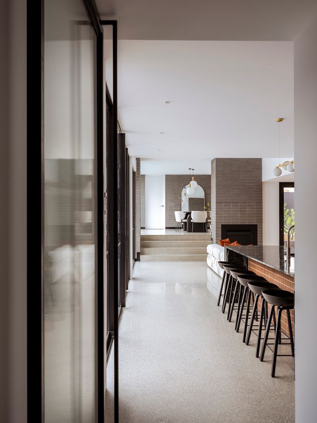 A long, bright contemporary open-plan interior viewed along a polished light terrazzo or concrete floor. The foreground contains a large island or bar with a dark benchtop, warm brick-tile front, and a row of black bar stools. A white curved sofa faces a built-in black fireplace set into a textured grey tiled wall. At the far end, a raised dining area includes a round table, chairs, pendant lights, and an arched mirror. Full-height black-framed glazing lines the left side, while concealed ceiling lighting and minimalist fixtures create a refined, spacious feel.