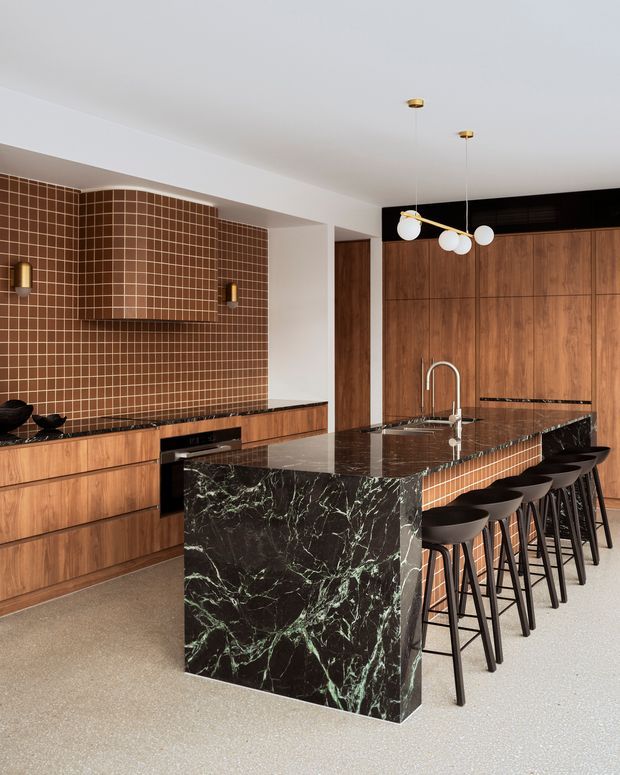 A sophisticated contemporary kitchen featuring extensive warm timber cabinetry, a curved brown square-tile backsplash and matching tiled island detail, a dramatic dark veined stone island, black accents, and a row of bar stools. Brass-and-white globe pendant lights hang above the island, while integrated appliances and a sculptural rangehood complete the refined, high-contrast interior.