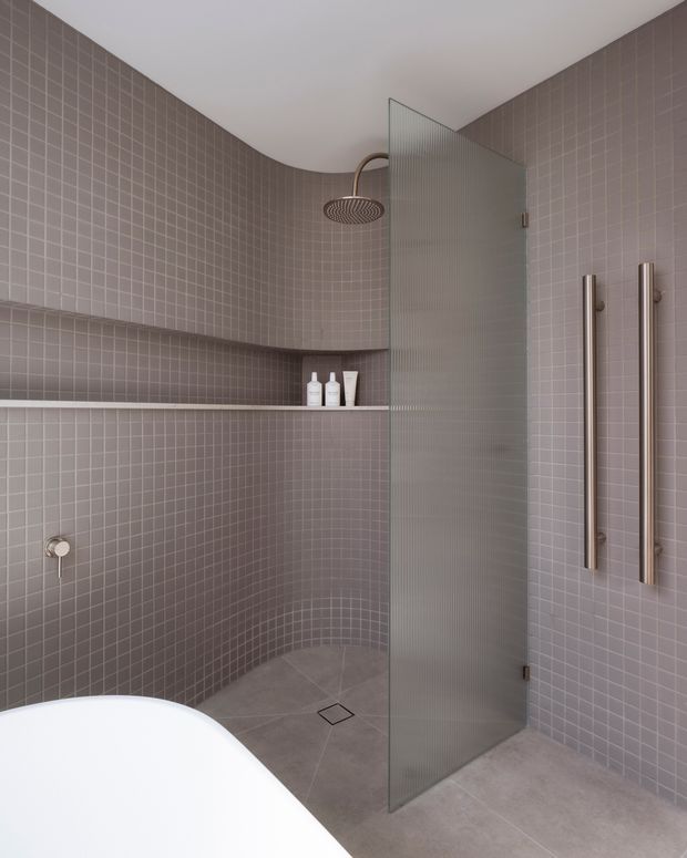 A contemporary bathroom featuring a curved walk-in shower lined with small muted mauve-grey mosaic tiles. A clear ribbed glass shower screen separates the shower from the room, with a round overhead shower, recessed tiled shelf, linear wall fixtures, and large-format grey floor tiles. The edge of a white freestanding bathtub is visible in the foreground.