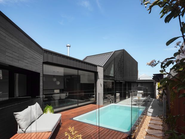 Contemporary dark-clad residential courtyard viewed from an elevated deck, featuring a long turquoise swimming pool enclosed by frameless glass balustrades, warm timber decking, a covered outdoor lounge, and large glazed openings into the house. The building has strong rectilinear volumes and a pitched metal roof with vertical screening. Bright daylight and contrasting dark architecture create a refined, modern atmosphere.