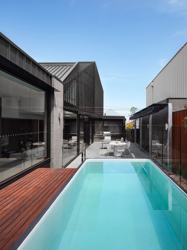A contemporary urban courtyard viewed from beside a narrow rectangular swimming pool. The space is enclosed by dark metal-clad and brick buildings with extensive floor-to-ceiling glazing, a timber pool deck, paved outdoor dining area, and a high slatted screening element. The restrained palette of charcoal, grey brick, glass, timber, and turquoise pool water creates a refined, modern architectural setting.