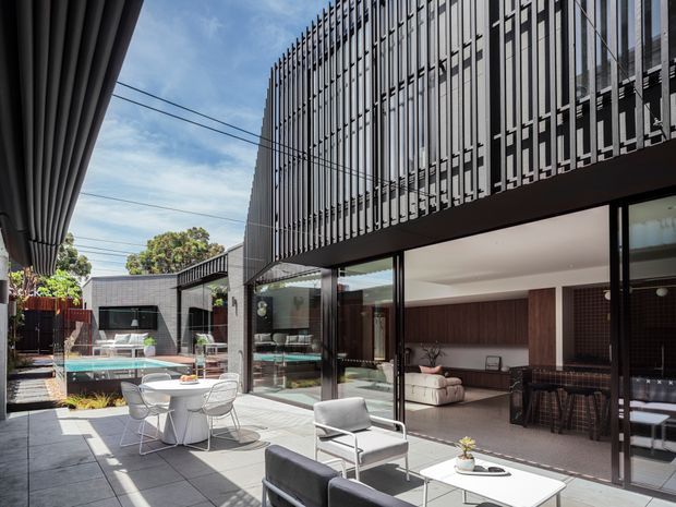 Contemporary multi-level residence viewed from a paved outdoor terrace, with strong vertical metal screening across the upper facade and extensive floor-to-ceiling glazing opening to a living and dining area. The terrace includes outdoor seating and a table, while a swimming pool and landscaped courtyard are visible beyond. The interior features dark timber wall panelling, a neutral sectional sofa, and a dining area with dark cabinetry or panelling.
