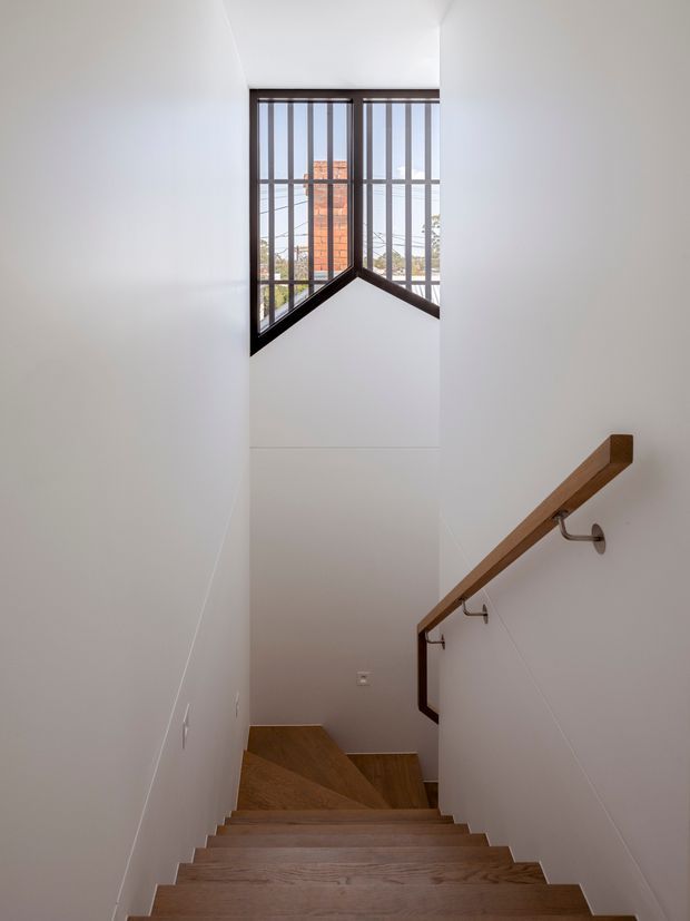 A narrow, bright stairwell viewed from above, with a flight of warm timber stairs descending toward a landing. White walls and ceiling create a clean, minimalist interior, while a timber handrail is mounted on the right wall with dark metal brackets. A tall angular window with dark framing and vertical security bars brings in daylight and reveals neighbouring brickwork outside.