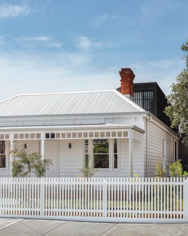 Street-facing view of a restored single-storey white weatherboard house with a corrugated metal gable roof, decorative verandah detailing, tall narrow windows, a red brick chimney, and a white picket fence. A dark contemporary upper-level extension is visible behind the traditional front portion.