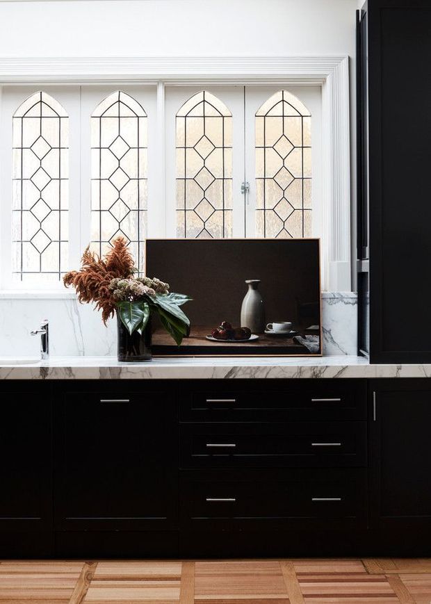 A refined kitchen or kitchenette with a dramatic black built-in cabinetry wall, white marble splashback and benchtop, and a central dark open niche styled with a vase, cups, and decorative objects. Tall arched leaded-glass windows with geometric diamond detailing and ornate white moulded trim bring light to the space. A timber parquet floor adds warmth and contrast.