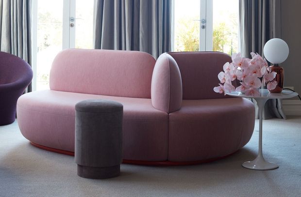 A refined contemporary living room featuring a sculptural blush-pink curved sofa with a dark red lower trim, a charcoal cylindrical ottoman, and a small sculptural side table topped with pink orchids and a spherical lamp. Floor-to-ceiling windows with layered grey curtains provide soft natural light, while the pale carpet and restrained furnishings create a polished, softly luxurious atmosphere.