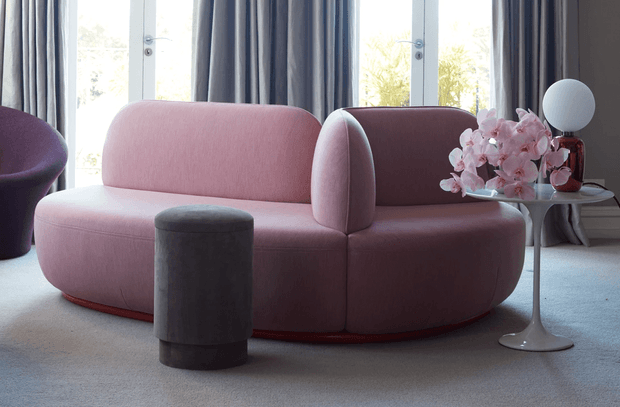 A stylish living room featuring a sculptural blush-pink curved sofa with rounded arms, a dark cylindrical ottoman, and a small sculptural side table holding pink flowers and a spherical lamp. Full-height windows with layered dark curtains provide soft natural light, while pale carpeting creates a calm, refined setting.