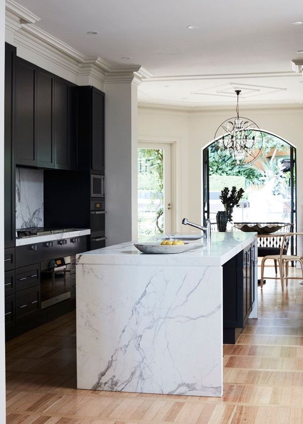 A refined kitchen with a large white veined stone island, dark charcoal-black cabinetry, integrated appliances, and a matching dark stone or marble splashback. The kitchen opens into a bright dining area through an arched glazed opening, with classical wall mouldings, decorative ceiling detailing, a sculptural chandelier, and warm timber parquet flooring.