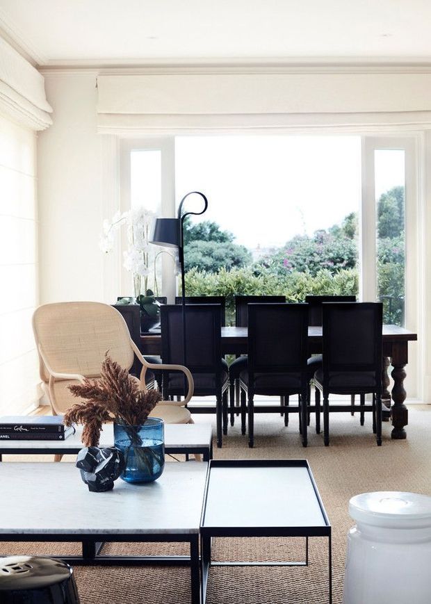 Bright, refined open-plan living and dining area with white panelled walls, decorative ceiling moulding, large windows overlooking a garden, a dark timber dining table and chairs, a pale lounge chair, and contemporary coffee tables with sculptural decor. The room has a classic architectural shell paired with modern furnishings.