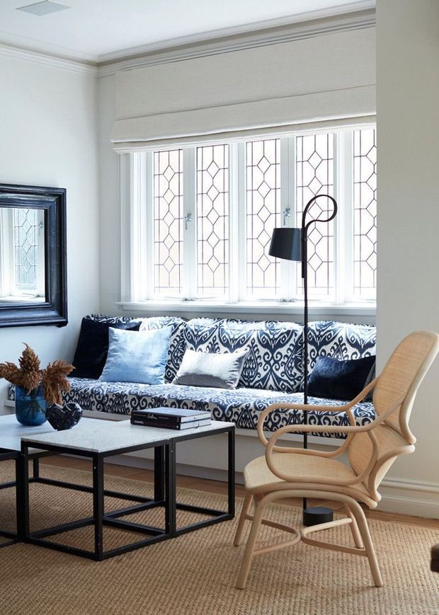 Bright, refined living room with a built-in window seat beneath tall patterned leaded-glass windows. The room features a blue-and-white patterned sofa, black-framed nesting coffee tables, a sculptural floor lamp, a woven rug, a cane lounge chair, framed mirror, and traditional crown moulding.
