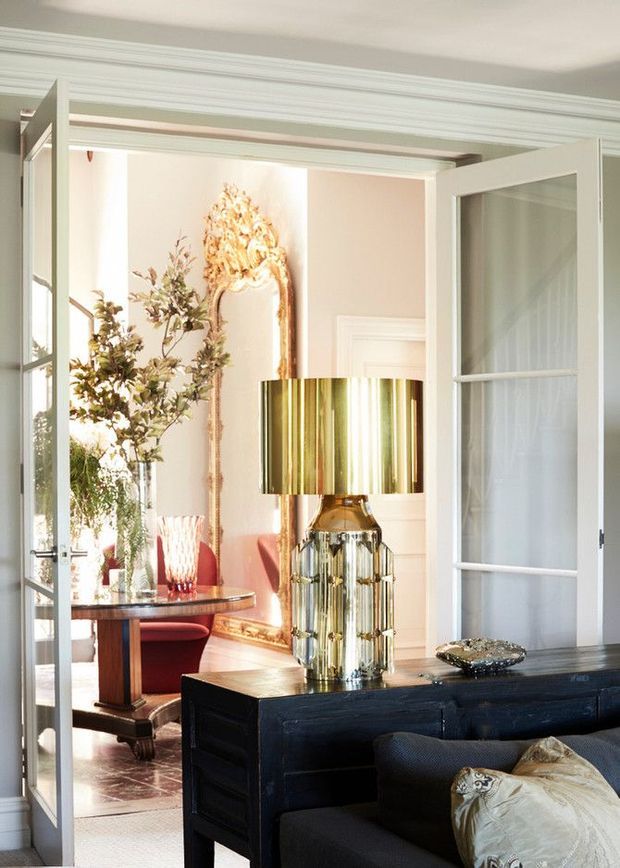 Elegant interior view through open glazed French doors, showing a refined living room in the foreground and a formal dining area beyond. The space features white traditional wall and door mouldings, pale blush walls, ornate gold-framed mirror, dark timber furniture, a polished metallic table lamp, red upholstered dining chairs, and decorative styling.