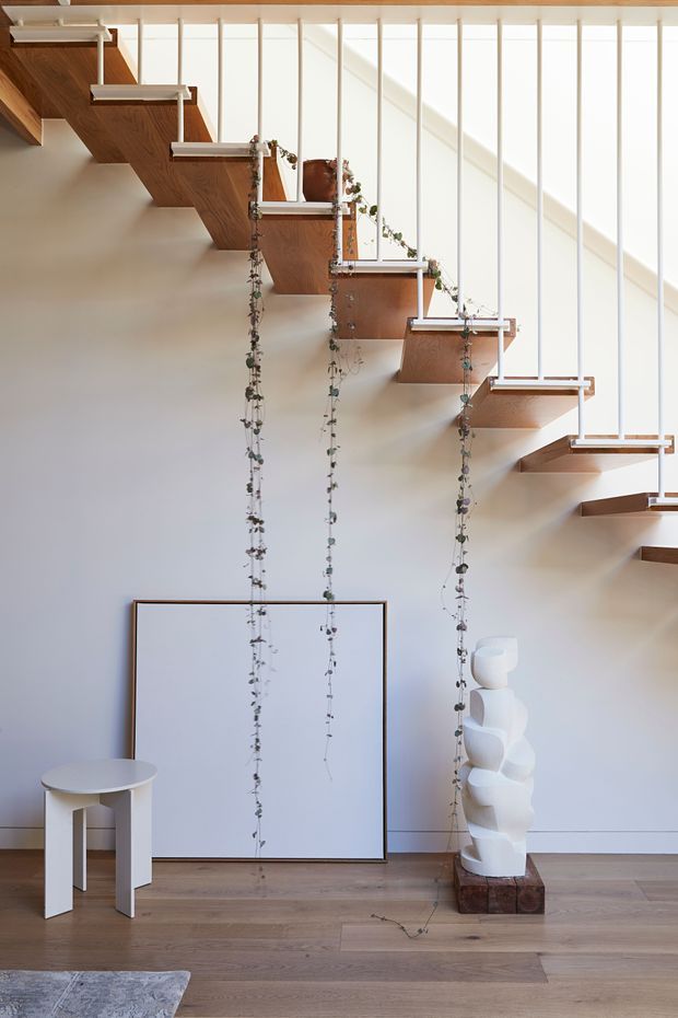 A bright, minimalist interior focused on a sculptural open staircase. Warm timber stair treads and landings project from a pale wall, supported by slender vertical white balusters and accented with trailing plants. The space has a light timber floor, white walls, sparse sculptural furnishings, and generous daylight from tall glazing or clerestory windows.