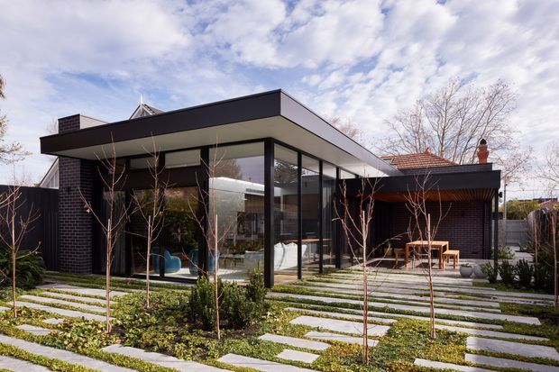 Exterior view of a contemporary single-storey home with a strong flat-roofed pavilion and extensive floor-to-ceiling glazing. Dark brick walls and dark window frames contrast with pale roof soffits, while a red-tiled roof section is visible behind the main volume. The foreground features a landscaped garden with regularly spaced stepping slabs and young deciduous trees, and a paved outdoor seating area is visible to the right.