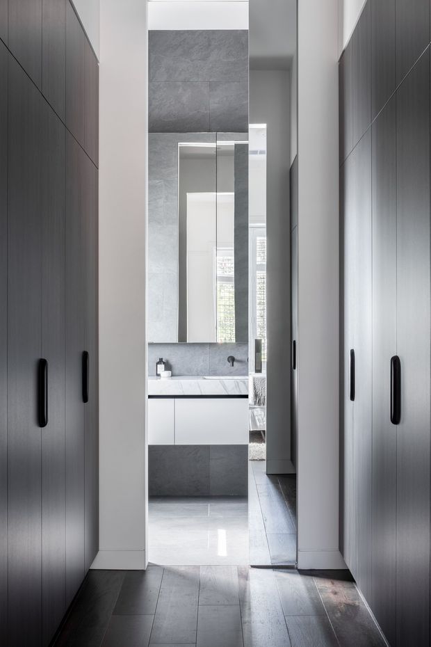A narrow, highly tailored hallway or dressing passage framed by tall dark built-in wardrobes with slim black handles. It opens directly into a compact contemporary bathroom featuring grey large-format tiled walls, a mirrored vanity wall, white floating cabinetry, and a pale stone or engineered-stone benchtop. Dark timber-look flooring continues through the passage, with bright natural light visible beyond.