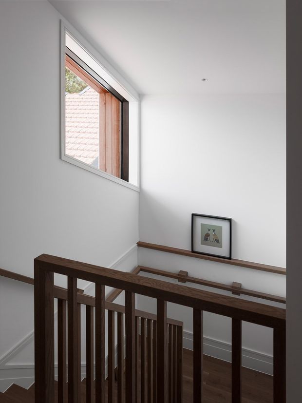 A minimalist residential stairway and upper landing viewed from above, with a dark timber balustrade and handrail, white painted walls, dark timber flooring, and a large high-set window. A framed artwork rests on the landing wall, while a secondary lower flight and matching timber handrail are visible below. Natural light creates a bright, quiet interior.