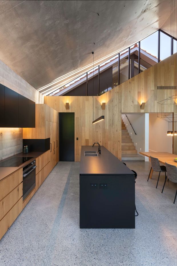 A sophisticated open-plan kitchen and dining area with a long matte-black island, integrated sink, timber cabinetry, dark appliances, and a terrazzo-like floor. Vertical timber wall panelling continues through the room and around a staircase, while a large concrete ceiling and extensive high-level glazing create a dramatic, light-filled interior. Minimal black pendant lights and wall sconces add a refined contemporary feel.