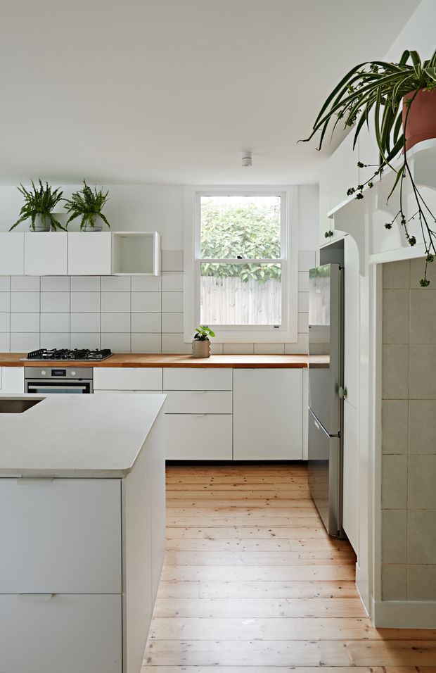 Bright, compact white kitchen with flat-panel cabinetry, a white tiled splashback, timber benchtops, and a large white island. A gas cooktop and built-in oven sit along the rear wall beneath open shelving and potted plants. Natural light enters through a central window, while pale timber floorboards and abundant greenery create a fresh, inviting feel. A tiled opening on the right leads toward another area, with a stainless-steel refrigerator beside it.