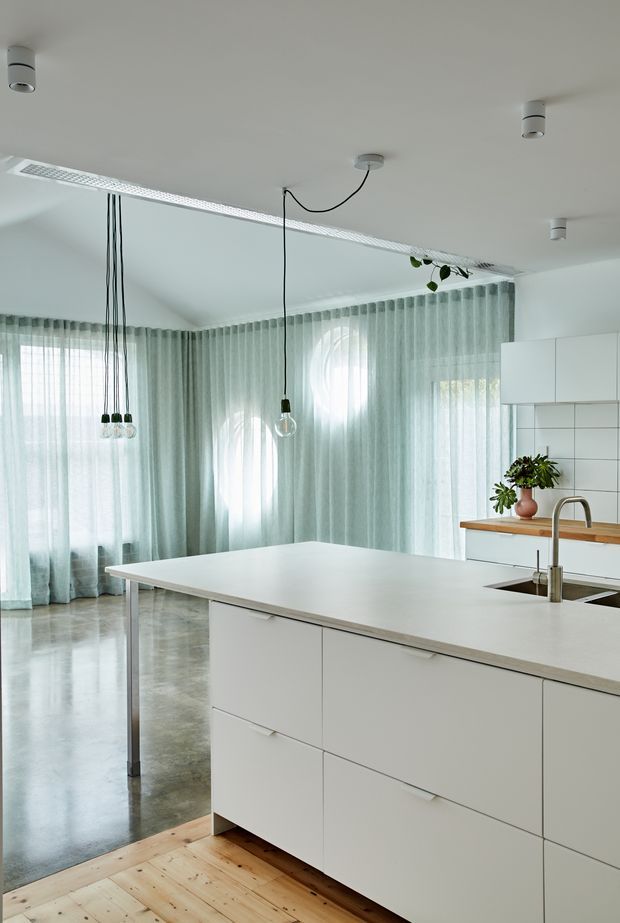 Bright, open-plan contemporary kitchen dominated by a large white island with integrated drawers, a stainless-steel tap and undermount sink. White flat-panel cabinetry lines the rear wall, paired with a warm timber benchtop and white tiled splashback. Floor-to-ceiling sheer pale curtains wrap the corner, with exposed pendant bulbs and simple ceiling downlights. The space combines polished concrete flooring with a small area of light timber flooring and abundant diffused daylight.