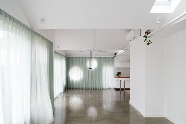 Bright open-plan interior with full-height pale sage-green sheer curtains, polished concrete flooring, white walls and ceiling, a minimalist pendant light, ceiling fan, skylight, and a partially visible compact kitchen with pale cabinetry and tiled splashback. The space feels airy and contemporary, with indoor planting and abundant diffused daylight.