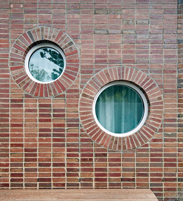 Close-up exterior view of a red and brown brick facade with two distinctive circular windows, each surrounded by radial brick voussoirs and pale trim. The lower edge shows a timber-toned horizontal ledge or deck element. Reflections of trees are visible in the upper window, while the larger lower window reveals sheer curtains.