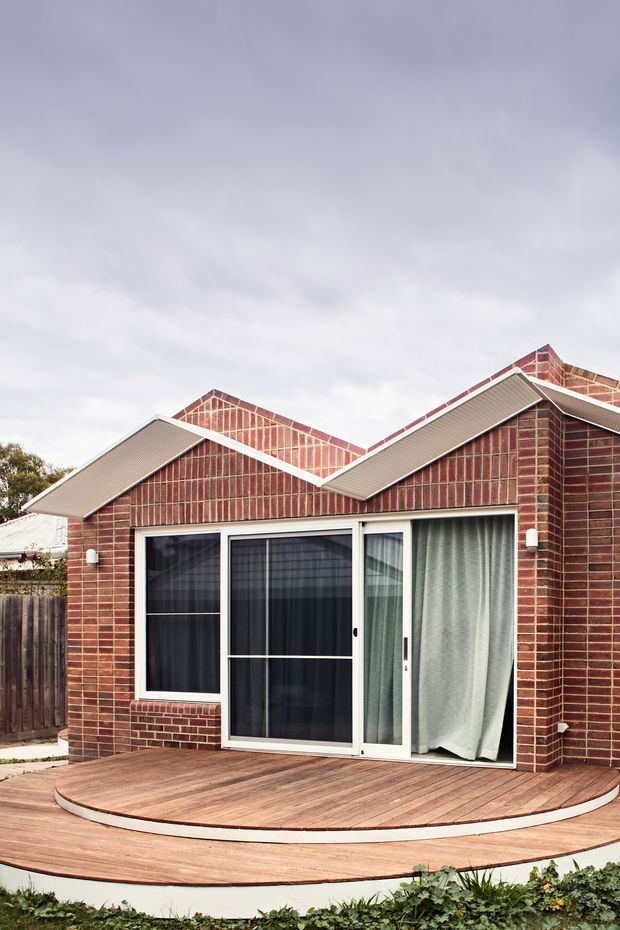 Exterior view of a compact red-brick residential extension with multiple pitched gables, white-framed glazed sliding doors, and a curved timber deck platform in front. The brickwork and roof forms dominate the facade, with a modest landscaped garden at the base.