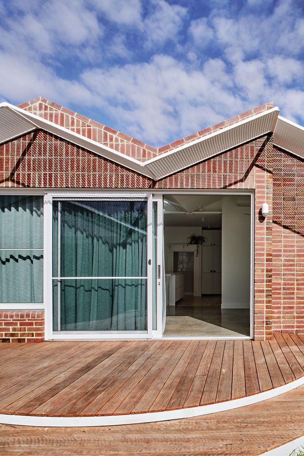 Exterior view of a brick house with a distinctive folded pitched roofline, white metal roof edging, and a timber deck curving across the foreground. Large glazed doors open directly onto the deck, revealing a simple interior with pale cabinetry and timber flooring. The image is taken in bright daylight beneath a partly cloudy blue sky.