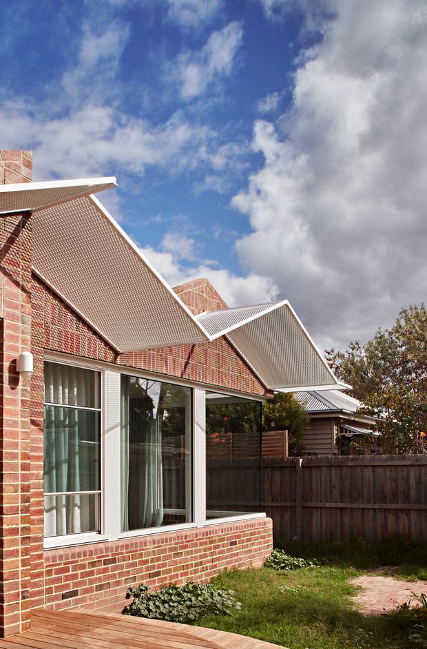 Exterior view of a brick residential extension with large glazed openings and distinctive corrugated metal skillion roof forms. The building sits beside a timber-fenced garden and deck under a partly cloudy blue sky.