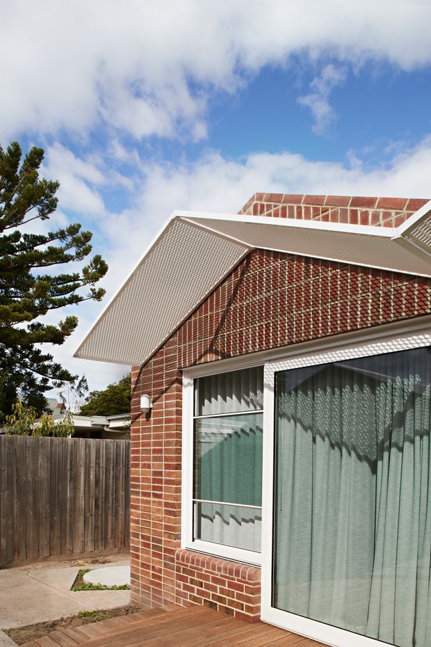 Close exterior view of a red brick residential building with a steep gabled roof and prominent white wire-mesh awnings or sunshades. White-framed windows and glazed doors reveal curtains inside. A timber deck, concrete path, timber boundary fence, and mature evergreen tree are visible around the house.