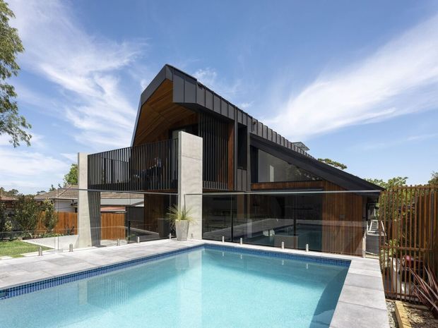 Exterior view of a contemporary multi-level residence with a dramatic sloping roof, dark vertical metal cladding, warm timber soffits and wall panels, concrete columns, extensive glazing, and a cantilevered upper level. A tiled swimming pool and paved terrace occupy the foreground, with a fenced suburban garden setting.