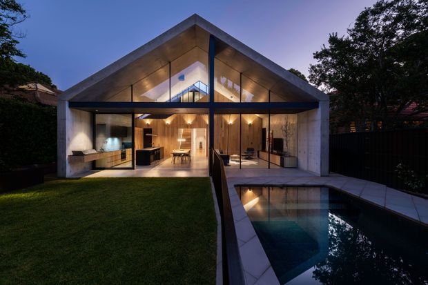 Twilight exterior view of a contemporary pavilion-like house with a steep gabled roof, exposed concrete frame, extensive floor-to-ceiling glazing, and warm timber-lined interiors. The home opens onto a lawn and a narrow swimming pool, with strong symmetrical geometry and illuminated interior spaces visible through the glass.