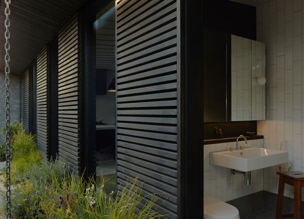 A moody contemporary bathroom viewed from a planted exterior or semi-enclosed walkway. The bathroom features a wall-mounted white rectangular basin, black vanity cabinetry and benchtop, white vertical rectangular wall tiles, a frameless mirrored cabinet, and a white toilet. Dark timber or painted horizontal louvred screens enclose the adjacent facade, with garden planting in the foreground.