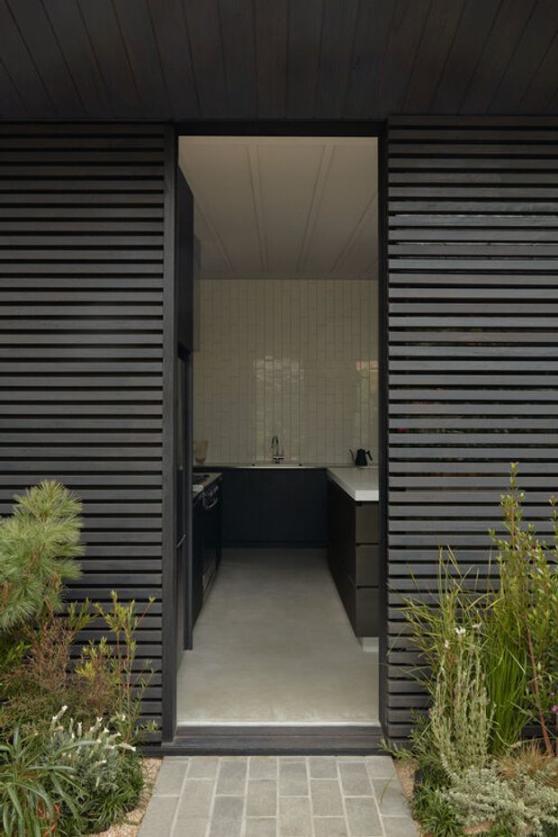 A dark, horizontally slatted exterior facade frames an open doorway into a compact contemporary kitchen. The interior features dark lower cabinetry, a pale stone or solid-surface benchtop, vertically patterned light tile walls, and a smooth concrete-like floor. A paved path and native-style planting lead to the entry, creating a restrained, modern indoor-outdoor composition.