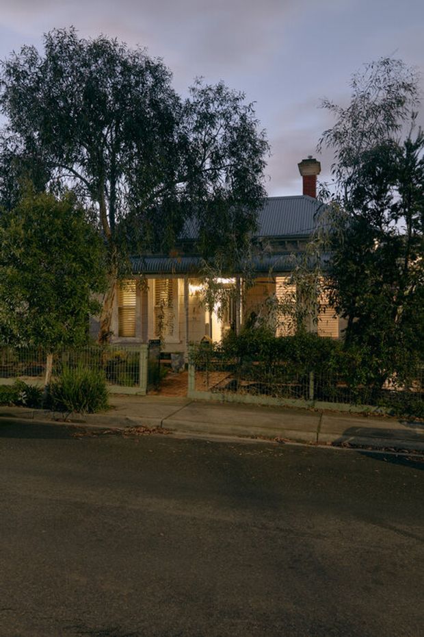 Dusk exterior view of a traditional single-storey weatherboard house set behind a leafy front garden and low metal fence. The home features a corrugated metal pitched roof, brick chimney, timber-framed windows and doors, and a small covered verandah with warm interior lighting. The camera is positioned across a residential street.