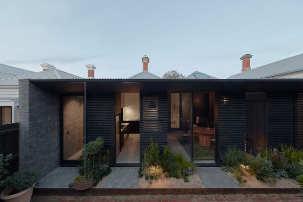 Front exterior view of a contemporary, dark-clad single-storey addition or townhouse facade. The building features a low-pitched roof with deep overhang, charcoal masonry, horizontal louvred screens, extensive dark-framed glazing, and a series of glazed entry doors. A narrow landscaped frontage with paving, gravel planting beds, grasses, and shrubs sits between the building and the street. Traditional neighbouring rooflines and chimneys are visible behind it.