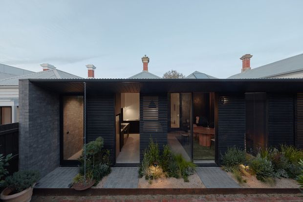 Single-storey contemporary residential facade viewed from the street, with a dark horizontal-roofed extension, charcoal masonry walls, black louvred screens, and floor-to-ceiling glazing. Warm interior lighting reveals an entry and living space behind the facade, while established neighbouring rooflines and chimneys sit in the background. A narrow landscaped frontage uses pavers, garden beds, and potted planting.