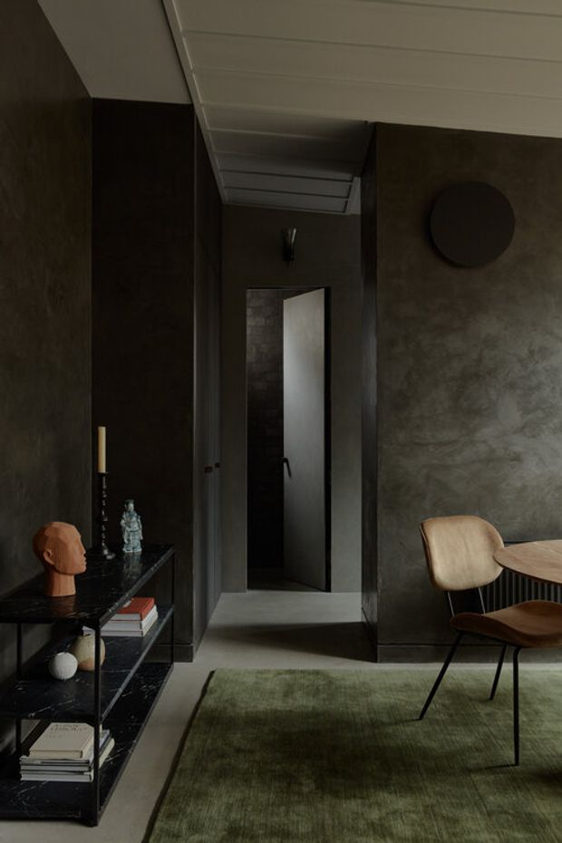 A moody, refined interior showing a dark-toned living or dining space. Textured charcoal walls, a white slatted ceiling, dark stone shelving, and a muted green rug frame a sculptural wooden dining chair and partially visible round table. A narrow hallway or doorway opens at the rear, while the shelving displays books and decorative objects.