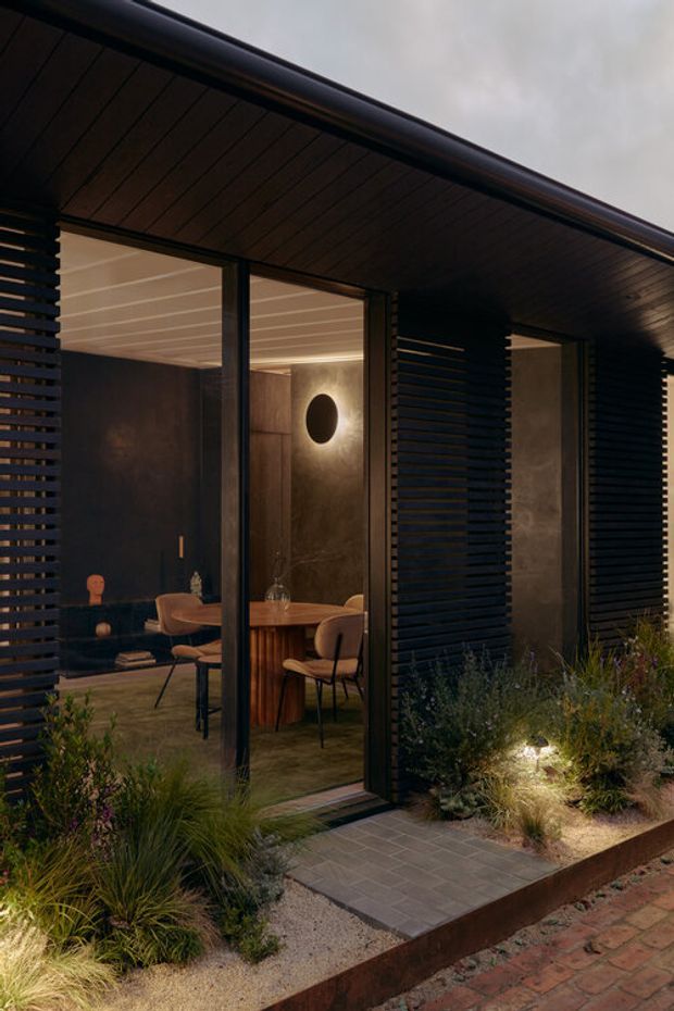 Exterior evening view of a dark, contemporary pavilion-like building with full-height glazed doors, deep timber-lined soffits, and horizontal dark slatted screening. The glazing reveals a warmly lit dining and lounge interior with a round wall light, timber dining table, upholstered chairs, and dark built-in seating. A paved entry path and layered drought-tolerant planting surround the building, with brick paving in the foreground.