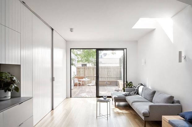 Bright contemporary living room with pale timber flooring, white walls, full-height vertical-panel built-in cabinetry, and a large grey sectional sofa. Black-framed glazed sliding doors open to a timber outdoor deck and fenced courtyard. A roof skylight brings strong natural light into the room, which has a clean, minimal fit-out.