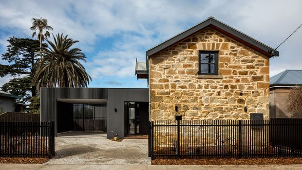Exterior view of a renovated stone dwelling combining a traditional gabled stone facade with a contemporary dark-clad addition. The property has a black metal picket fence, a wide paved driveway or entry forecourt, dark-framed glazing, and a corrugated metal roof. Mature palm and evergreen trees frame the streetscape.